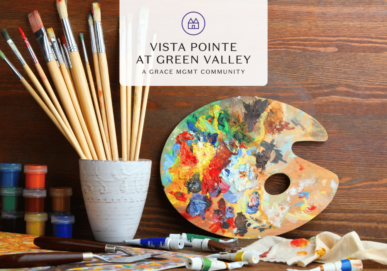 Artist's workspace with paintbrushes in a holder, colorful palette, and tubes of paint on a wooden table.