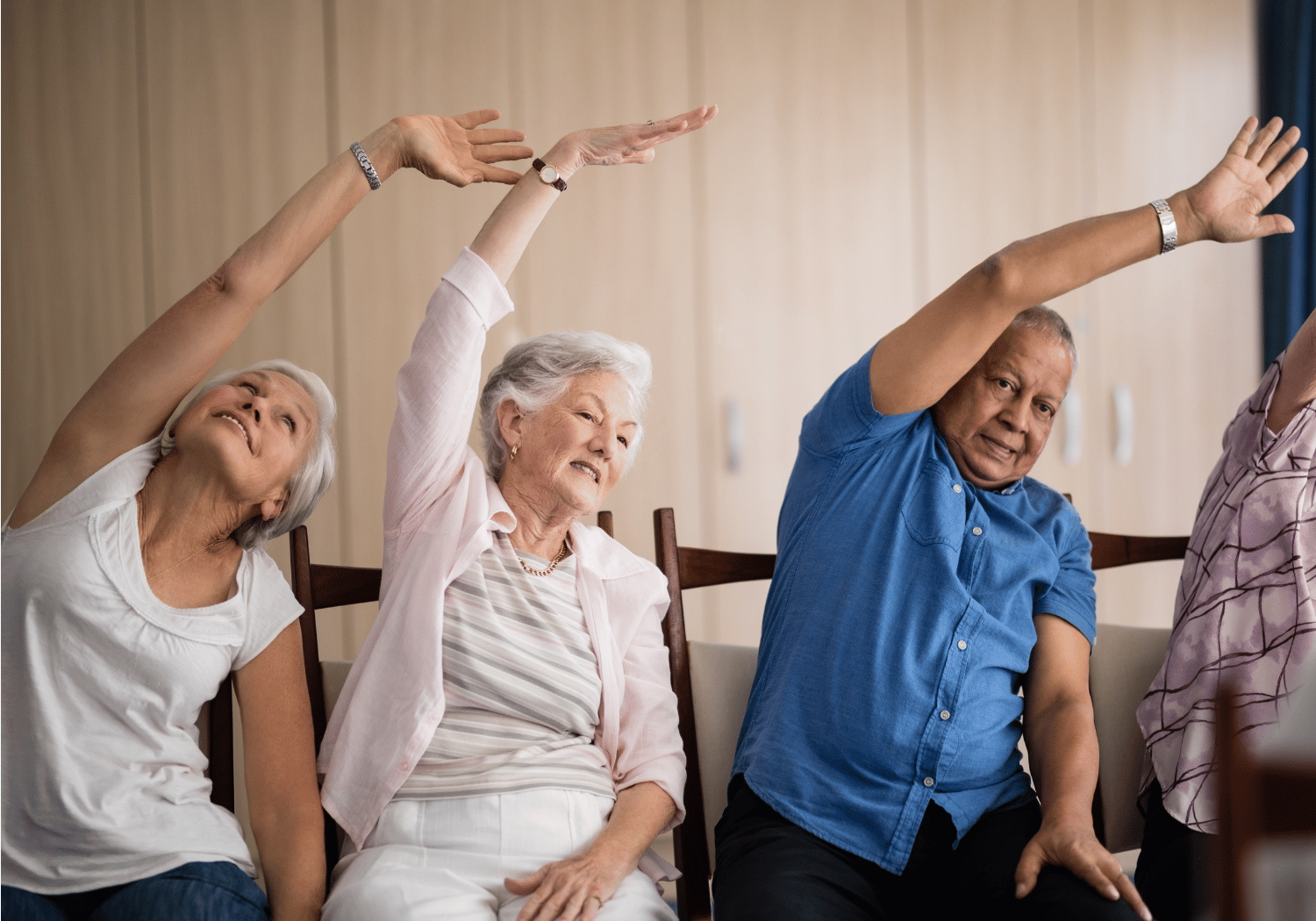 Four older adults seated in a row, raising arms overhead in a group stretch indoors takeaway: a gentle exercise class scene.