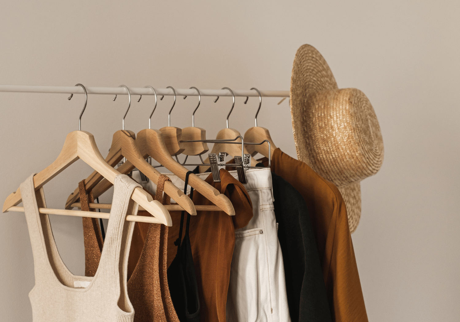 Wardrobe rail with neutral-toned clothes on wooden hangers and a woven straw hat on the end.
